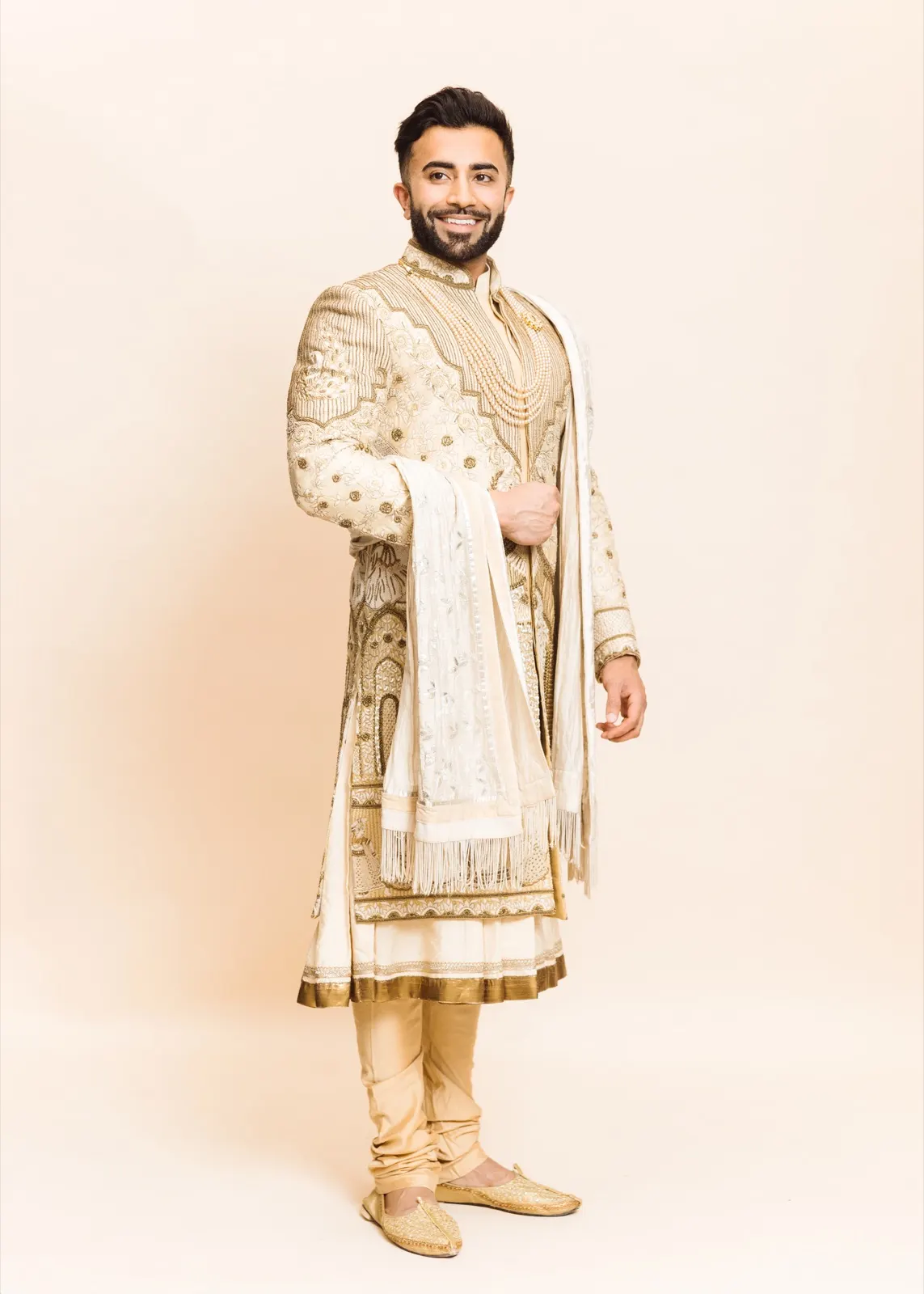 Hemel stands smiling in a full-length cream-and-gold sherwani with pearl necklaces and a flowing stole.