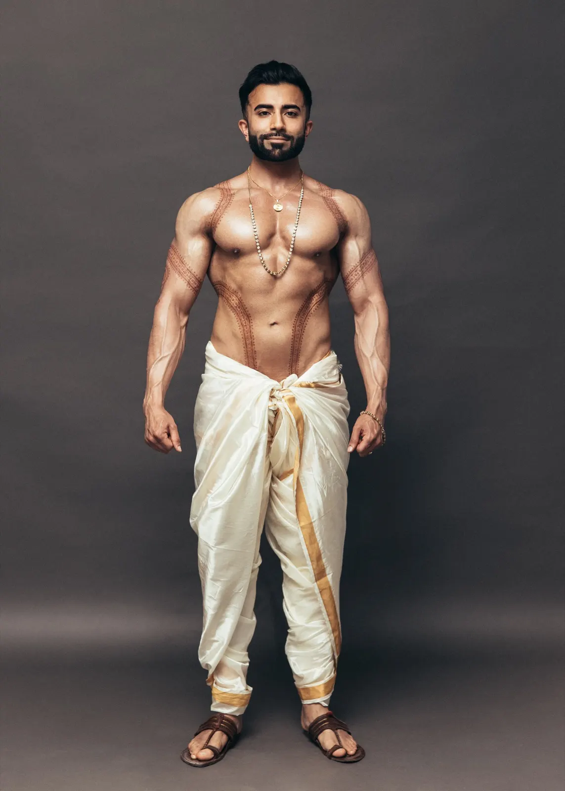 Hemel stands front-facing in an ivory-and-gold dhoti with jewellery and symmetrical traditional body art.