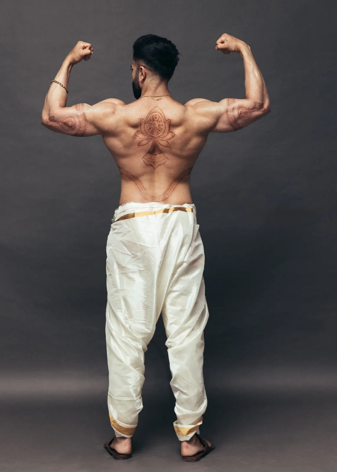 Hemel flexes from behind in an ivory dhoti, displaying detailed body art across his back and arms.