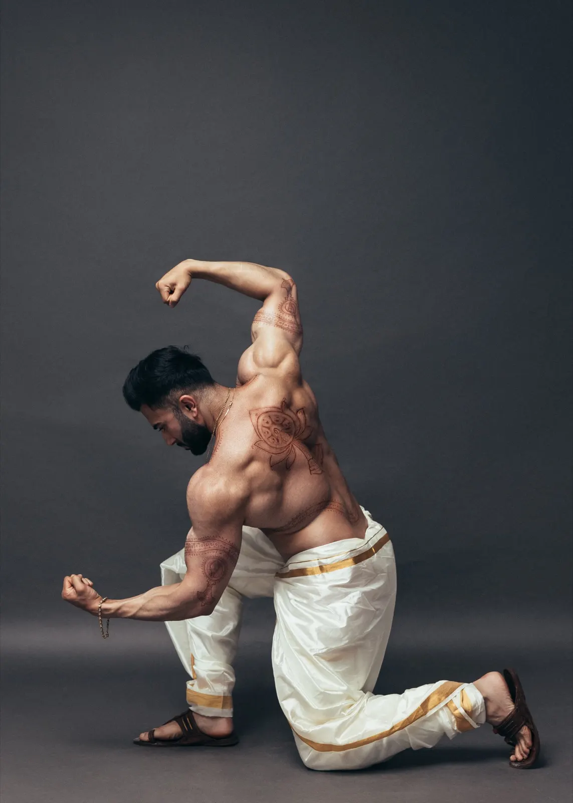 Hemel kneels in a dramatic dance-inspired pose wearing an ivory dhoti with traditional body art visible.