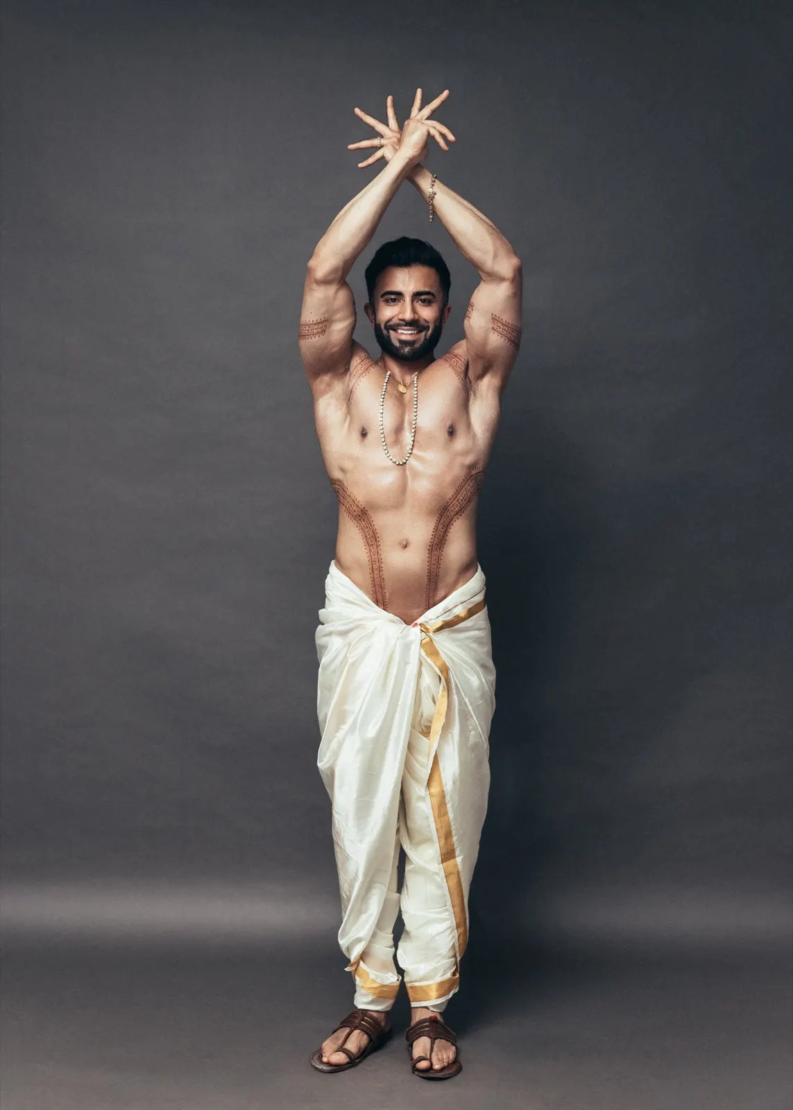 Hemel smiles with his arms raised in a symmetrical full-length pose wearing an ivory-and-gold dhoti.