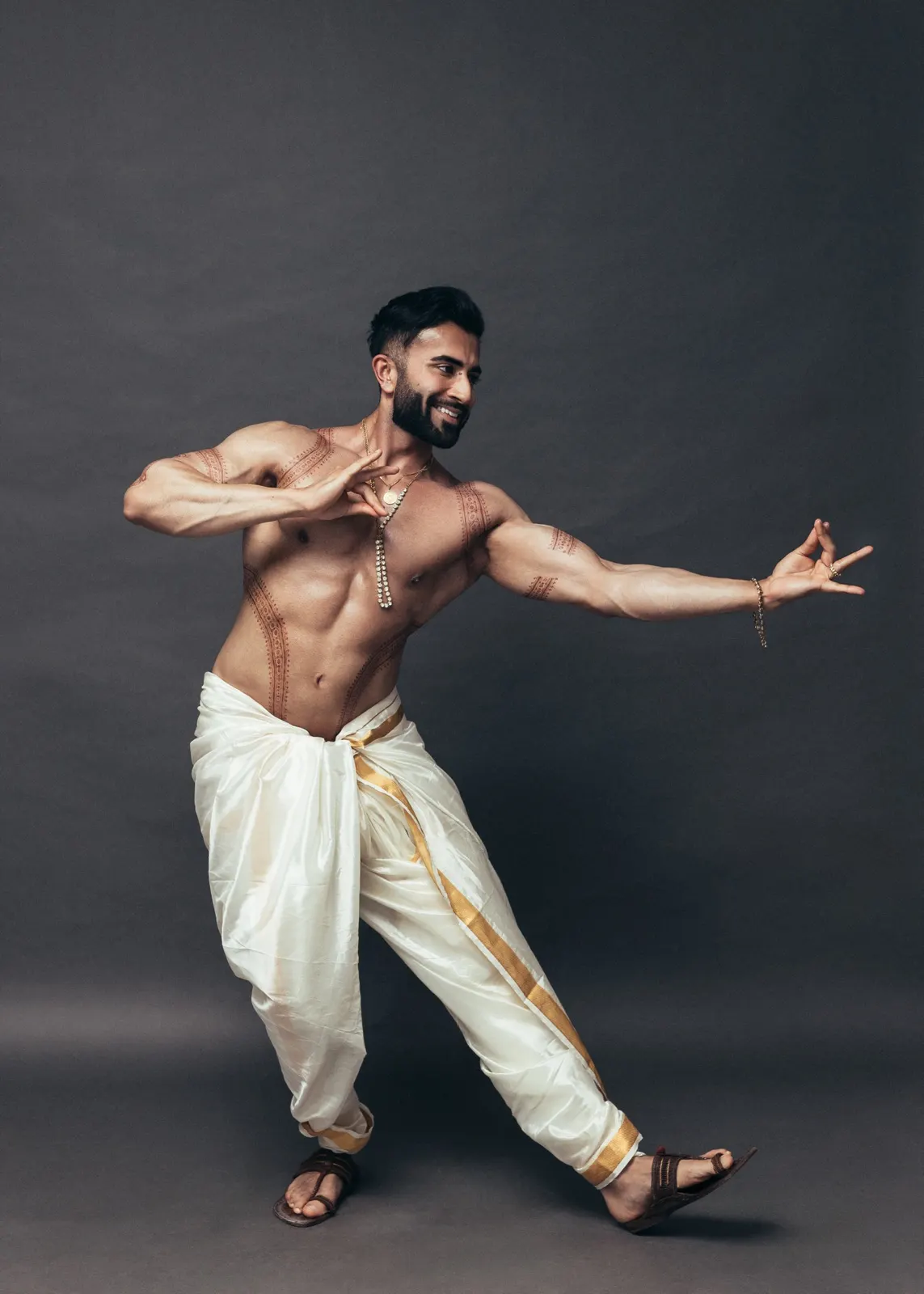 Hemel performs an expressive dance pose in an ivory-and-gold dhoti against a charcoal backdrop.