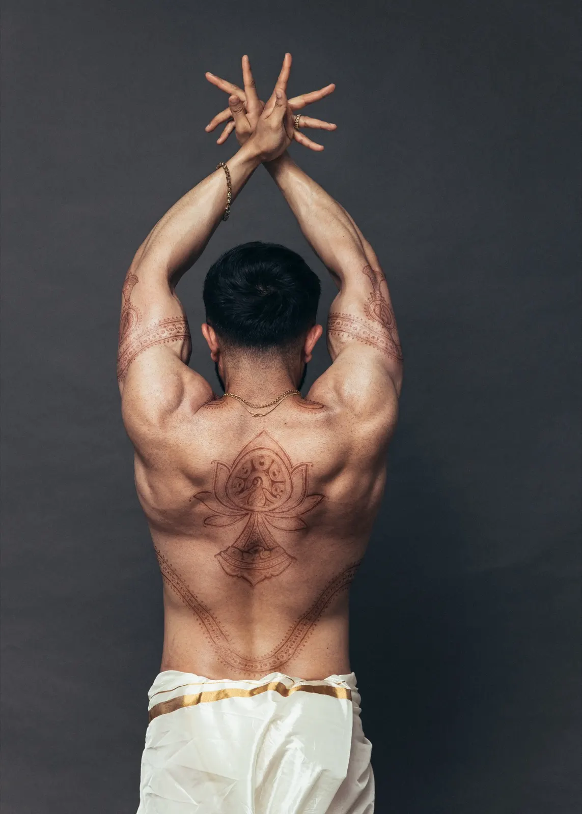 Hemel faces away with his arms raised, revealing symmetrical body art above an ivory dhoti.