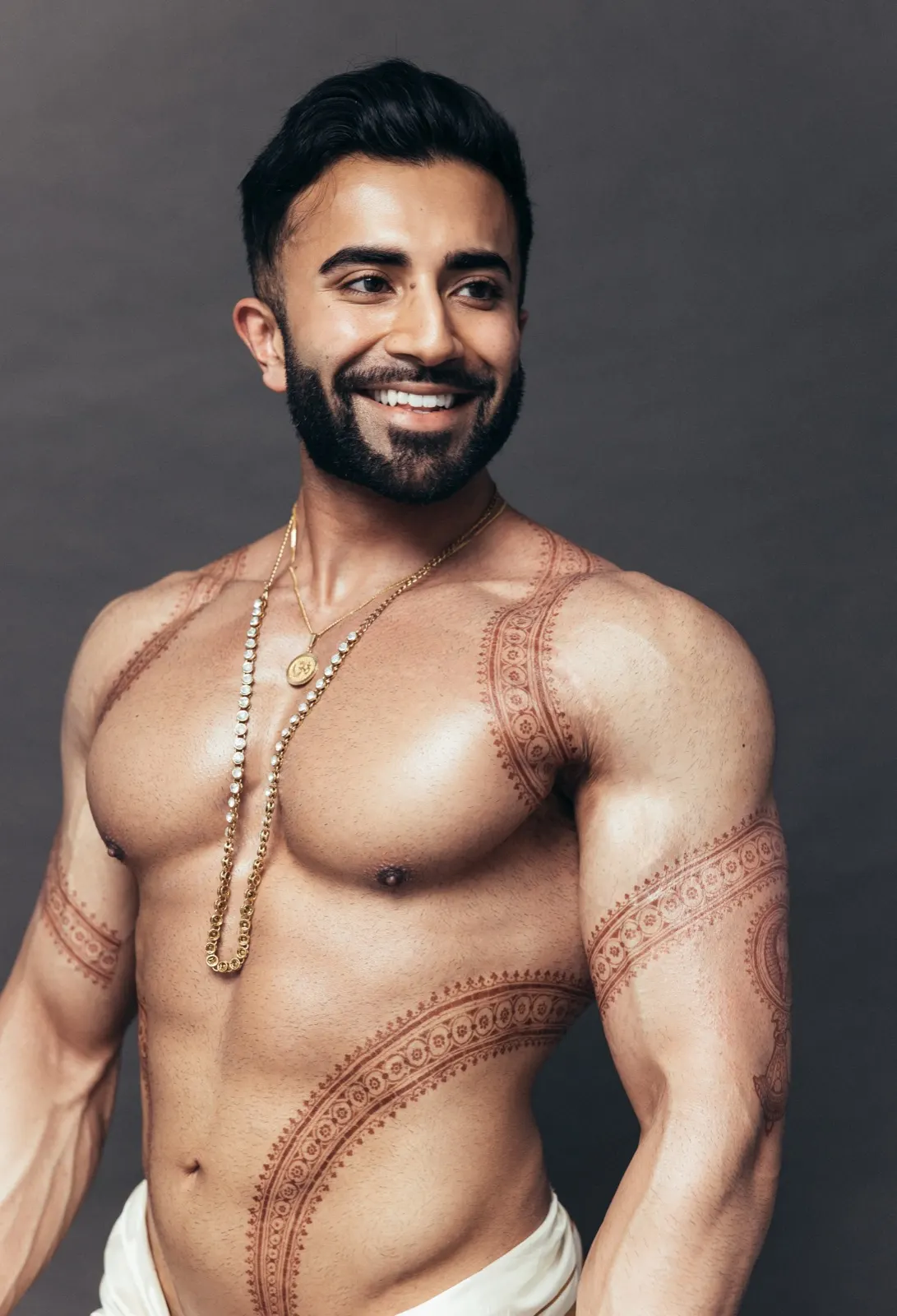 Hemel smiles in a close editorial portrait featuring gold necklaces and intricate traditional body art.