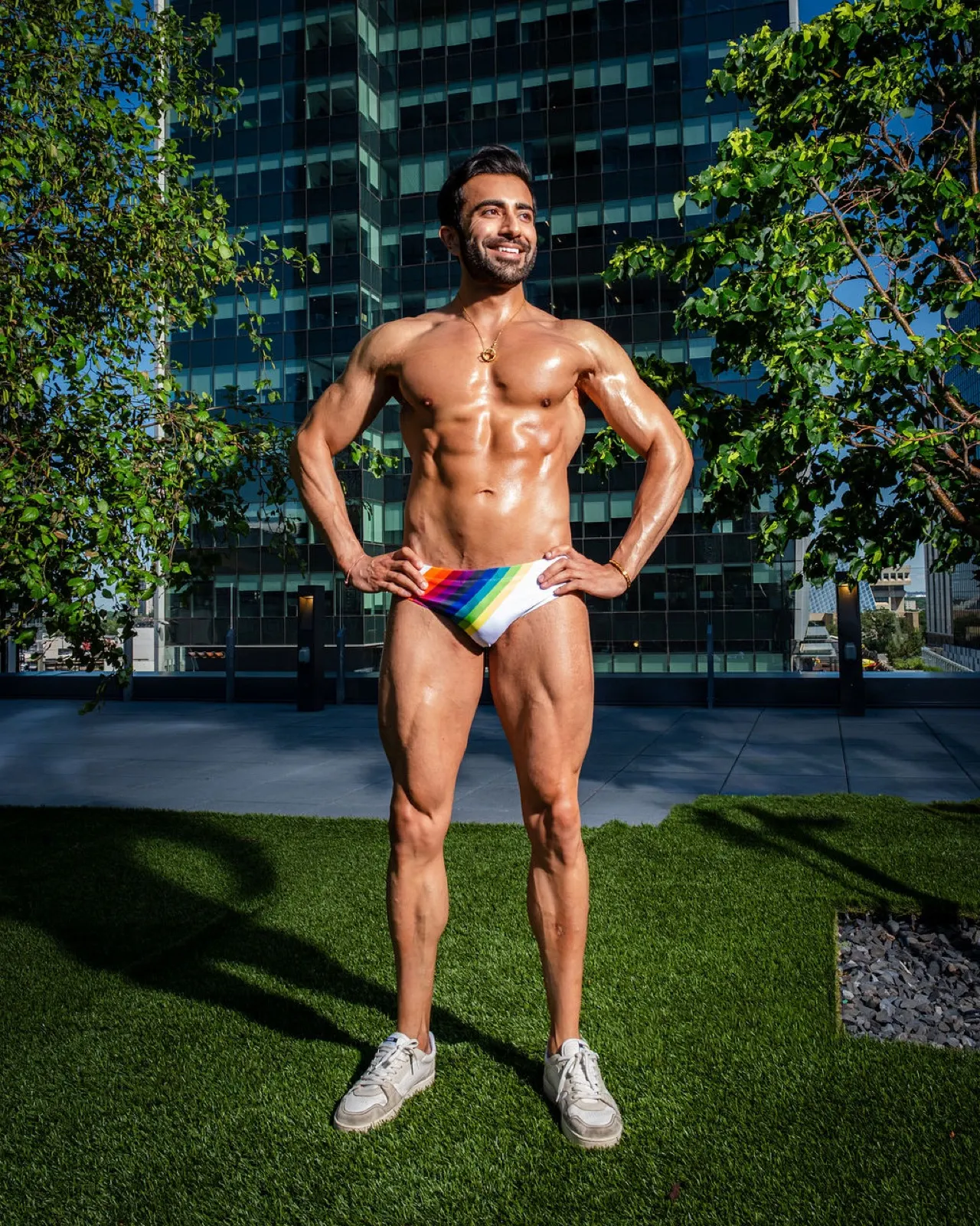 Hemel stands with hands on hips in rainbow swim briefs, surrounded by greenery and reflective city architecture.