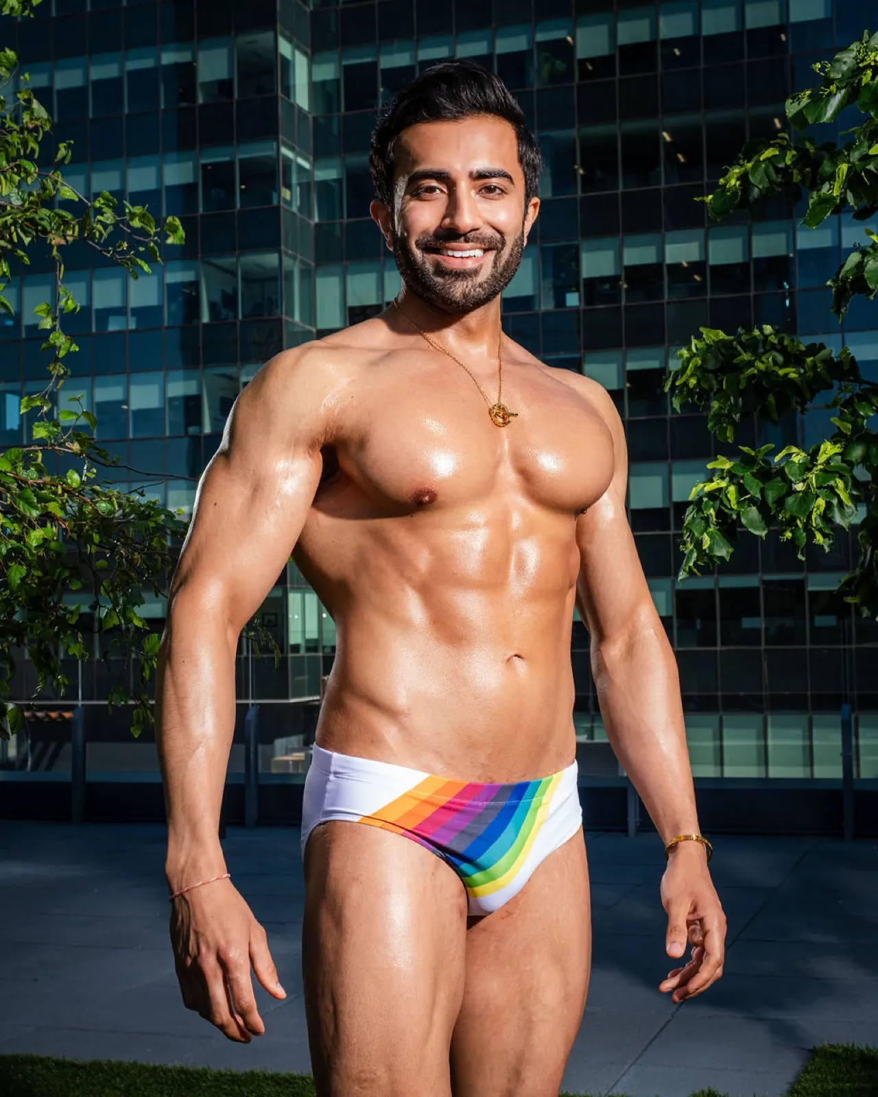 Hemel smiles confidently in rainbow swim briefs with glass towers and greenery behind him.