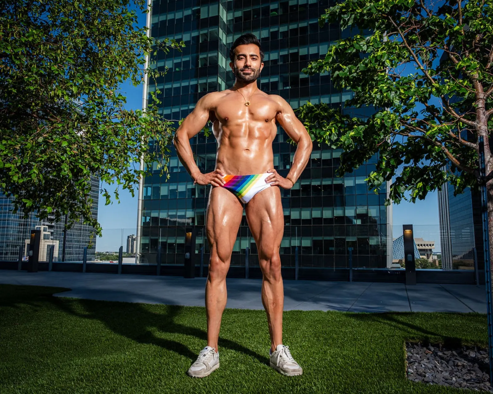 Hemel stands full length in rainbow swim briefs and sneakers, framed by trees and a glass office tower.