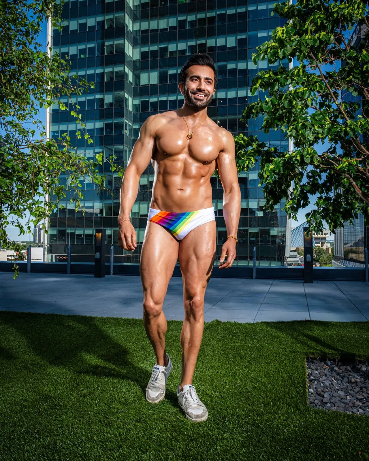 Hemel walks forward across a rooftop lawn in rainbow swim briefs with trees and glass buildings behind him.