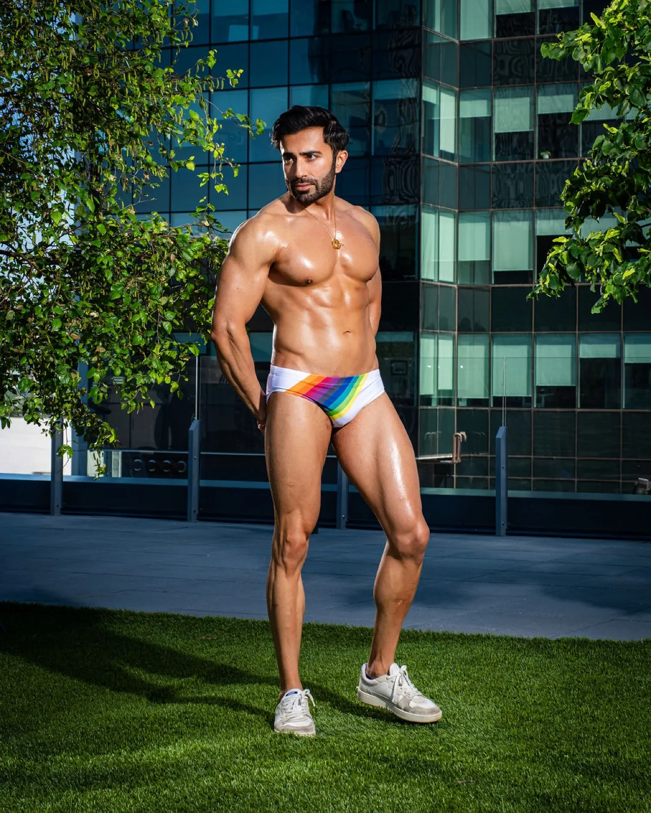 Hemel poses in profile with a focused expression, wearing rainbow swim briefs in front of a glass building.