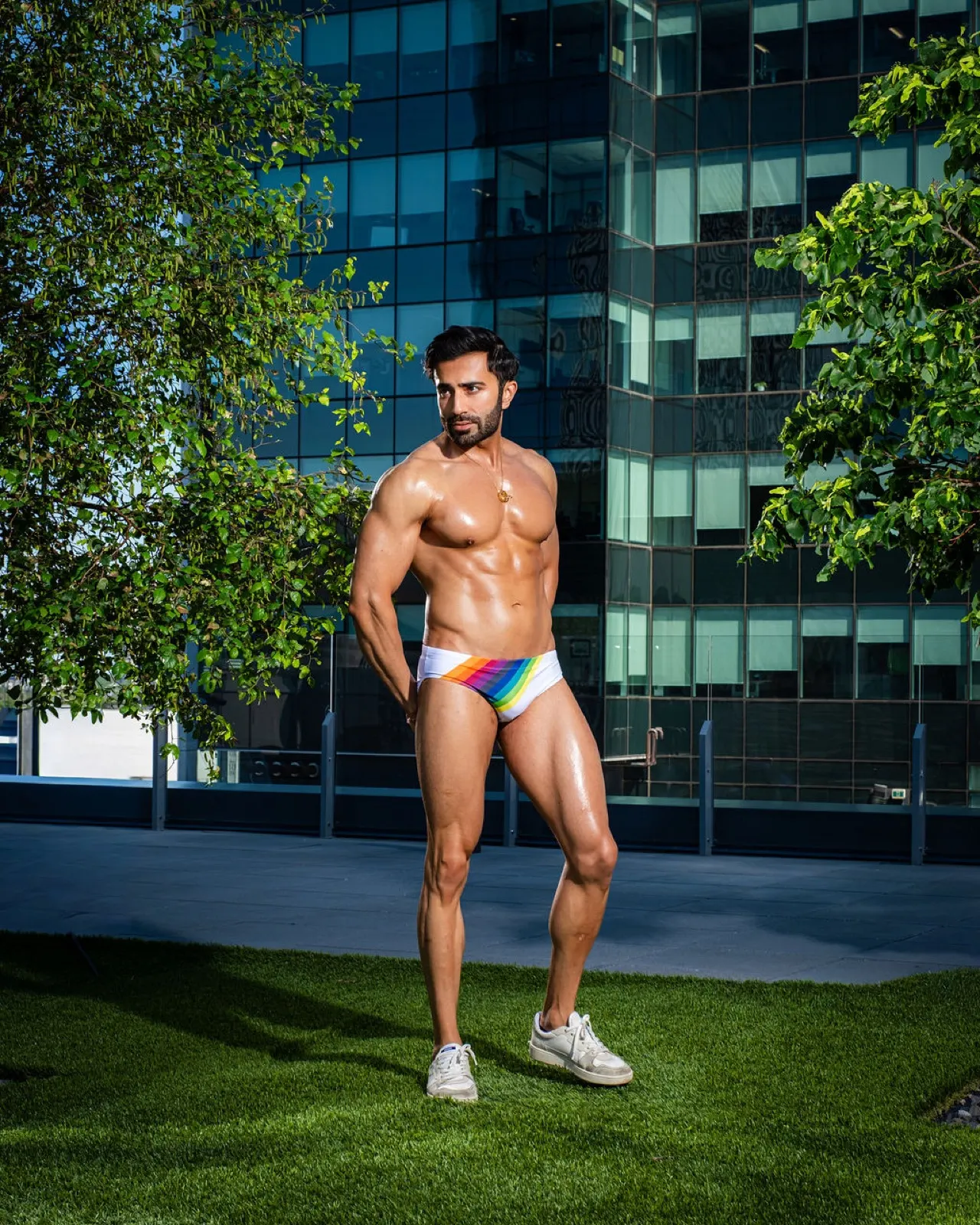 Hemel poses in rainbow swim briefs on a rooftop lawn, looking to the side between leafy trees and modern buildings.