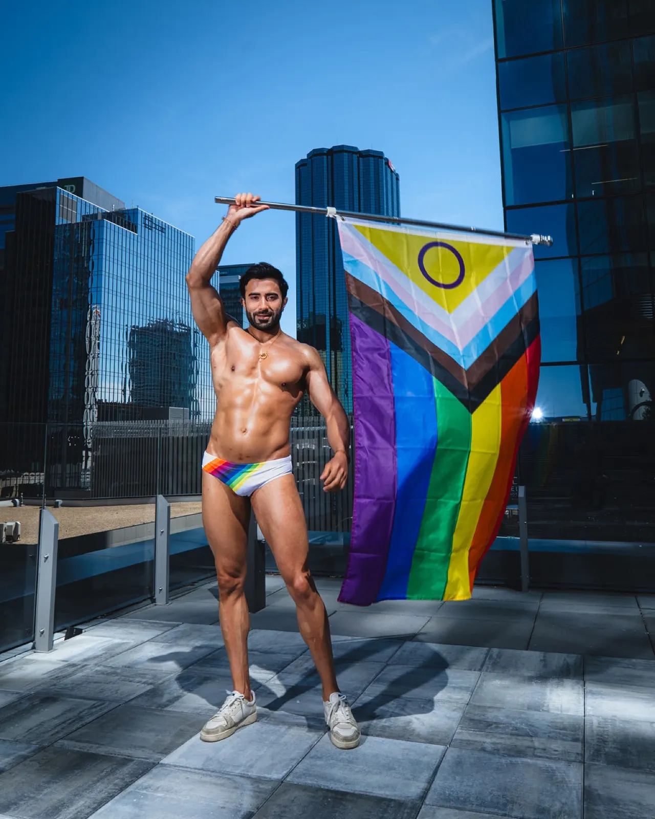 Hemel raises a Progress Pride flag on a city rooftop while wearing rainbow swim briefs.