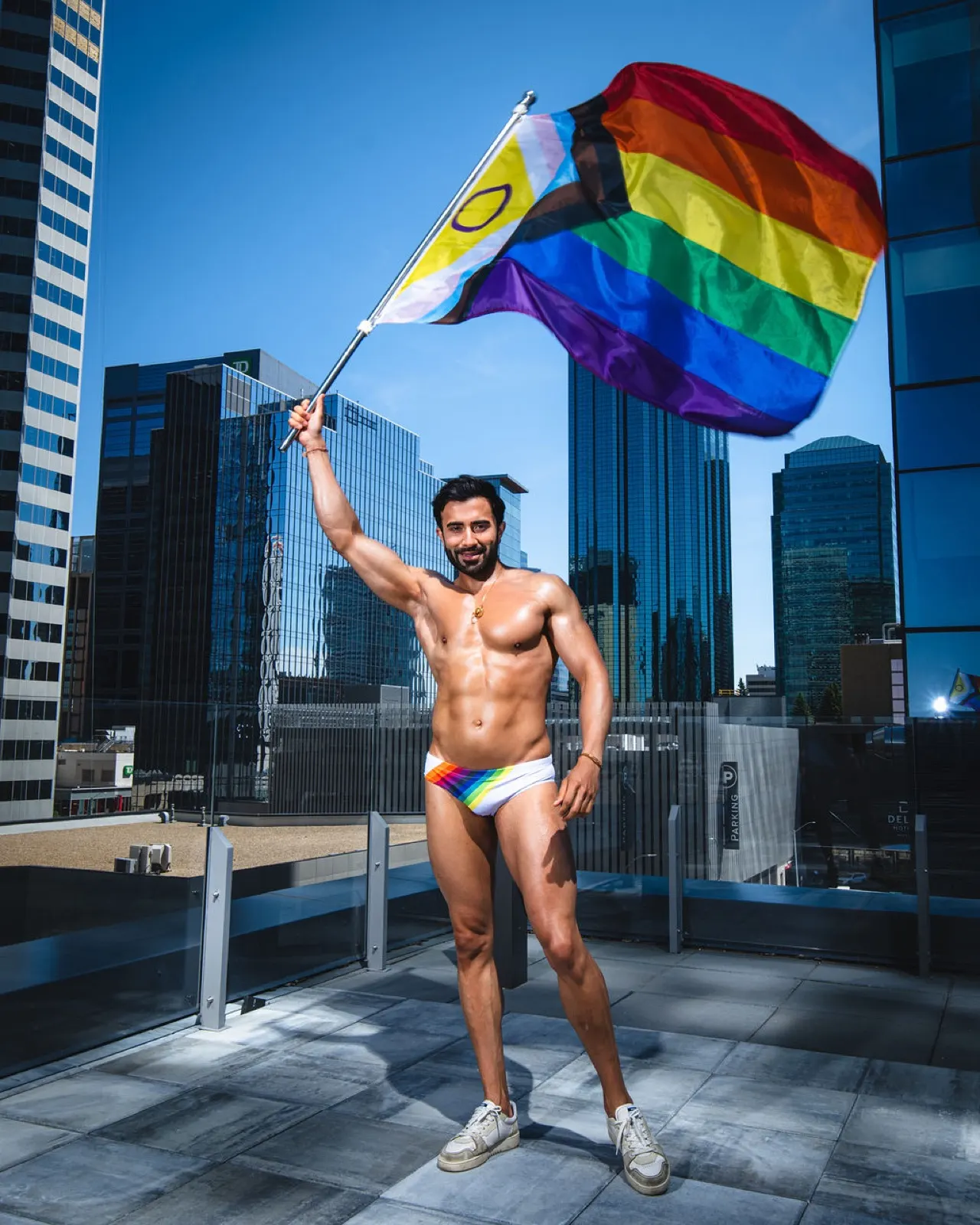 Hemel stands on a rooftop in Edmonton holding a large Pride flag above him against a downtown skyline.