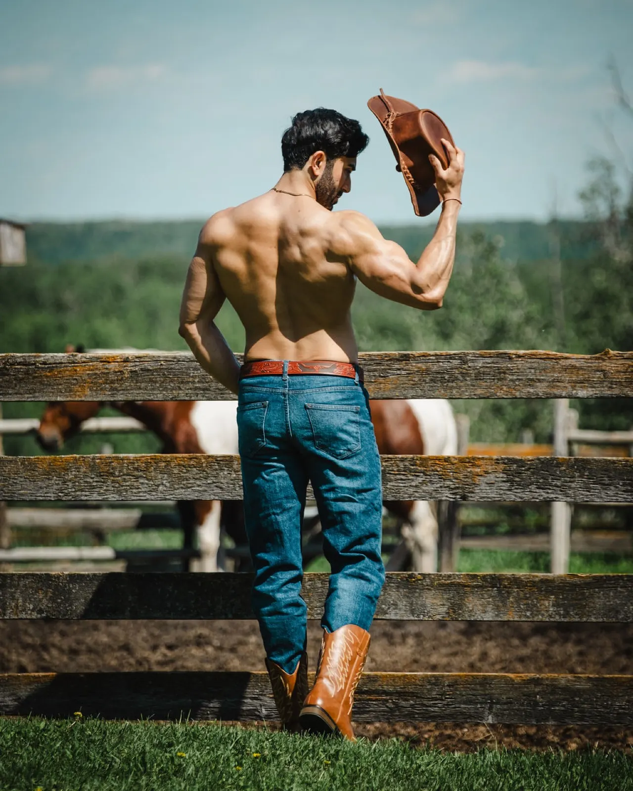 Hemel faces away by a wooden fence, showing a strong back silhouette while lifting a cowboy hat.