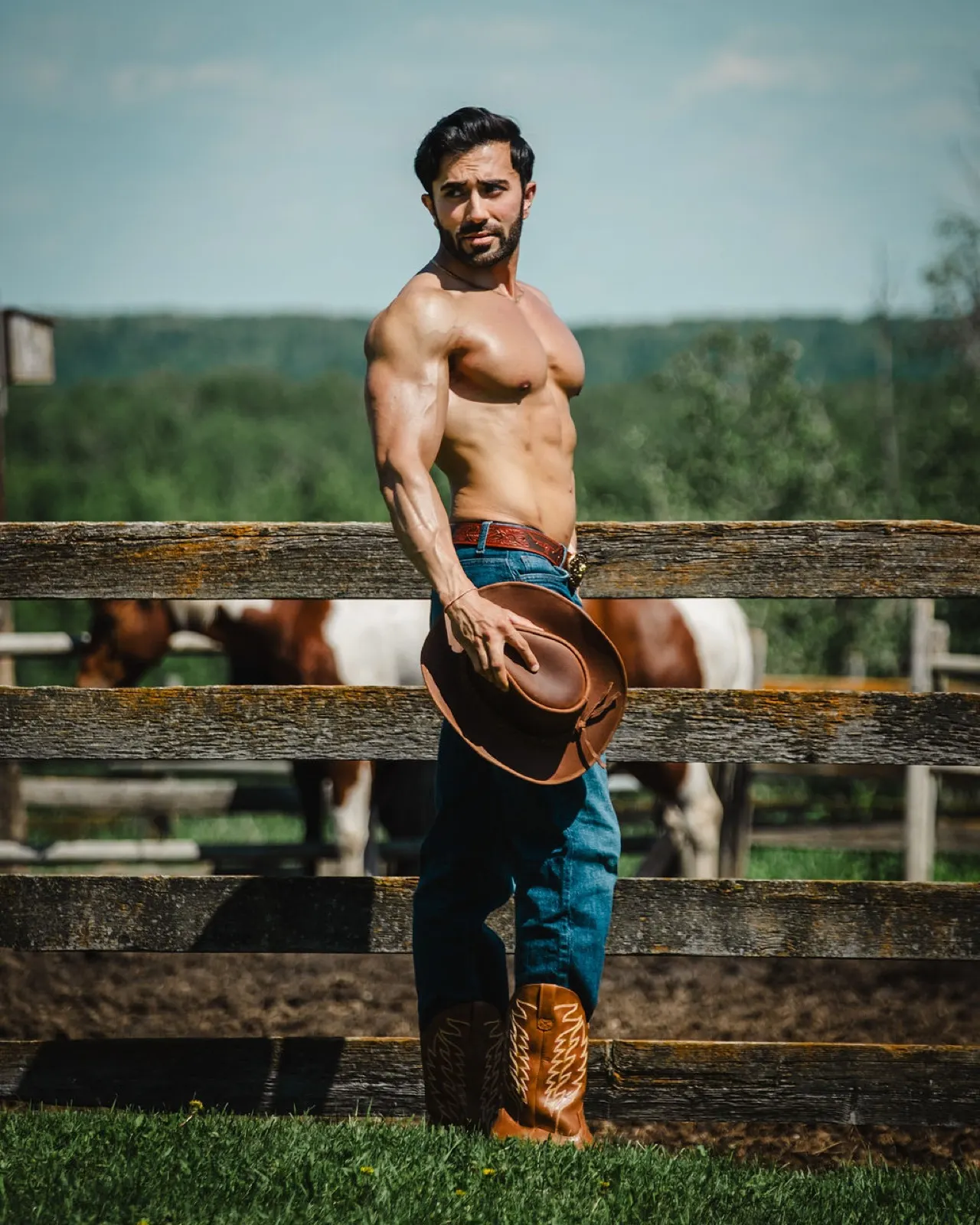Hemel turns toward the camera beside a wooden fence, holding a cowboy hat with horses behind him.