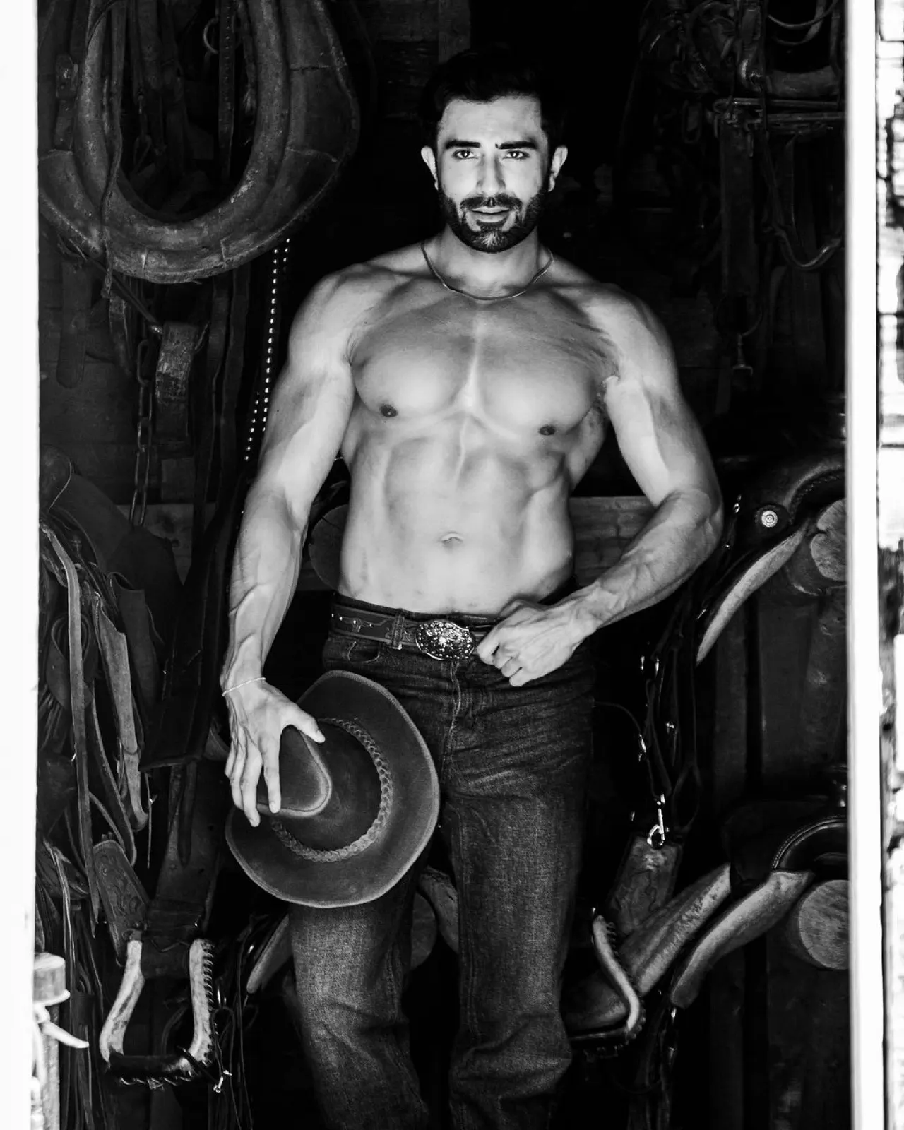Hemel poses in black and white inside a tack room, holding a cowboy hat among saddles and straps.