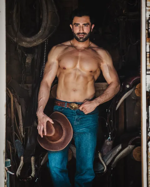 Hemel stands shirtless in a rustic saddle room holding a cowboy hat, styled in denim and western accessories.