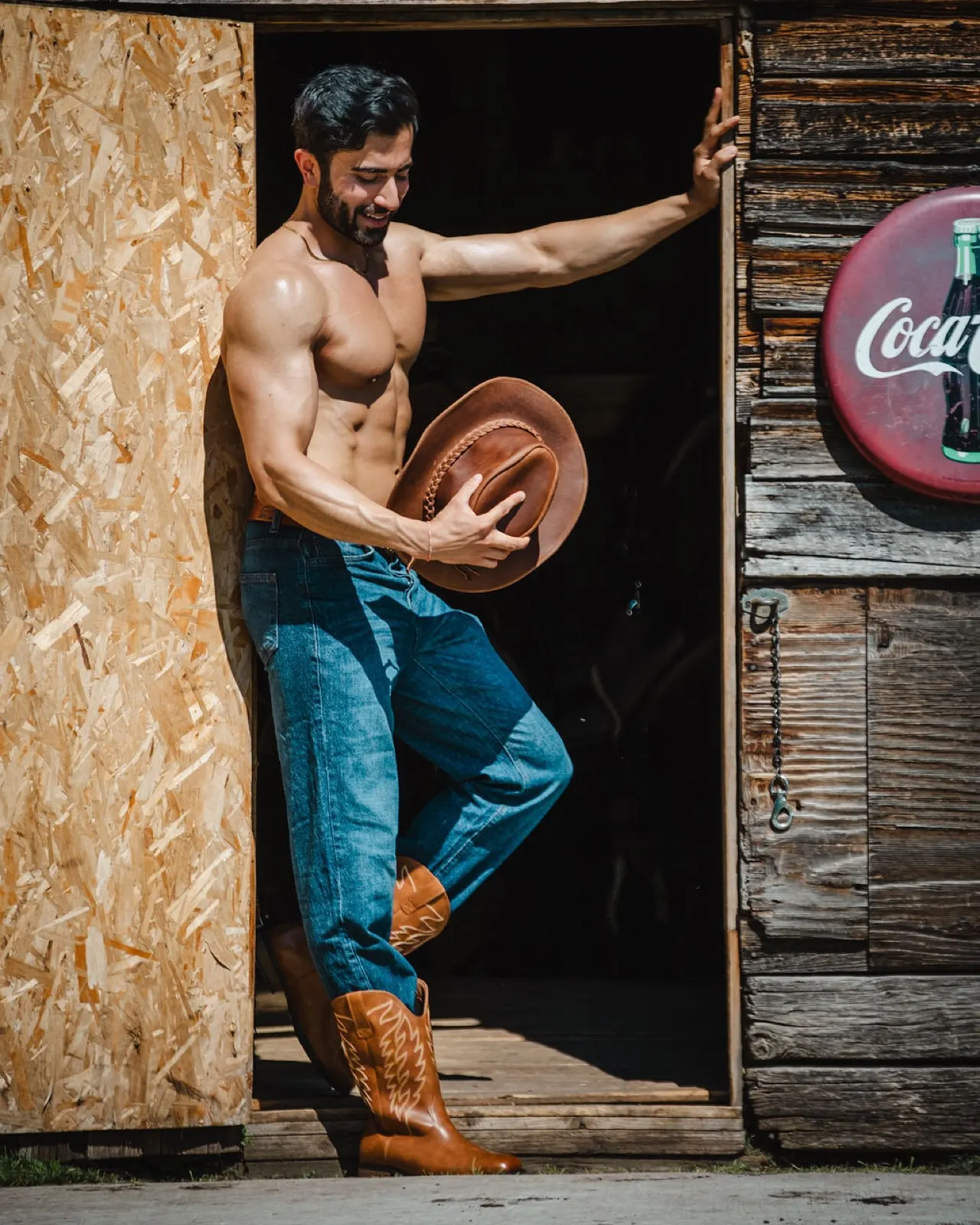 Hemel steps from a rustic barn doorway holding a cowboy hat, dressed in denim and boots.