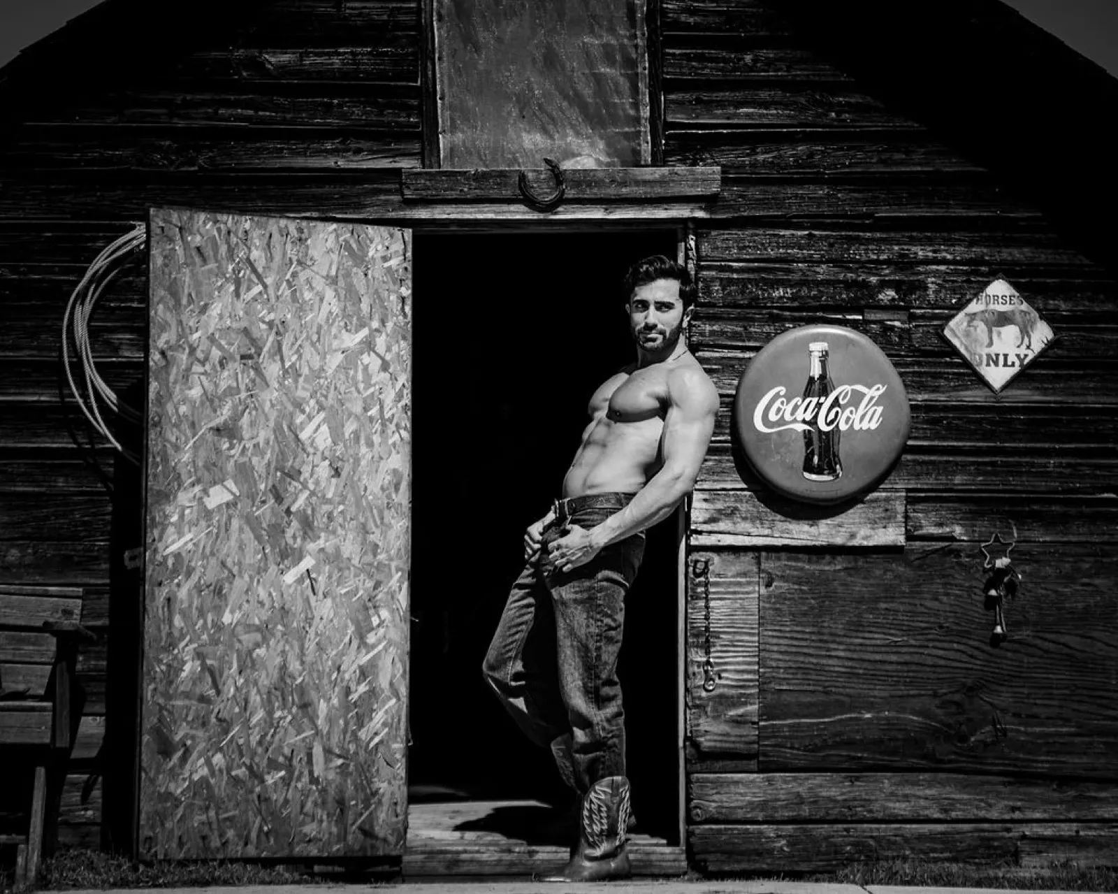 Hemel leans in a barn doorway in a black and white western scene with rustic signage.