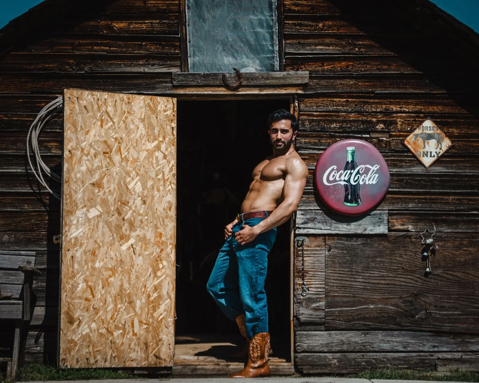 Hemel poses confidently in a barn doorway beside weathered wood and a vintage Coca-Cola sign.