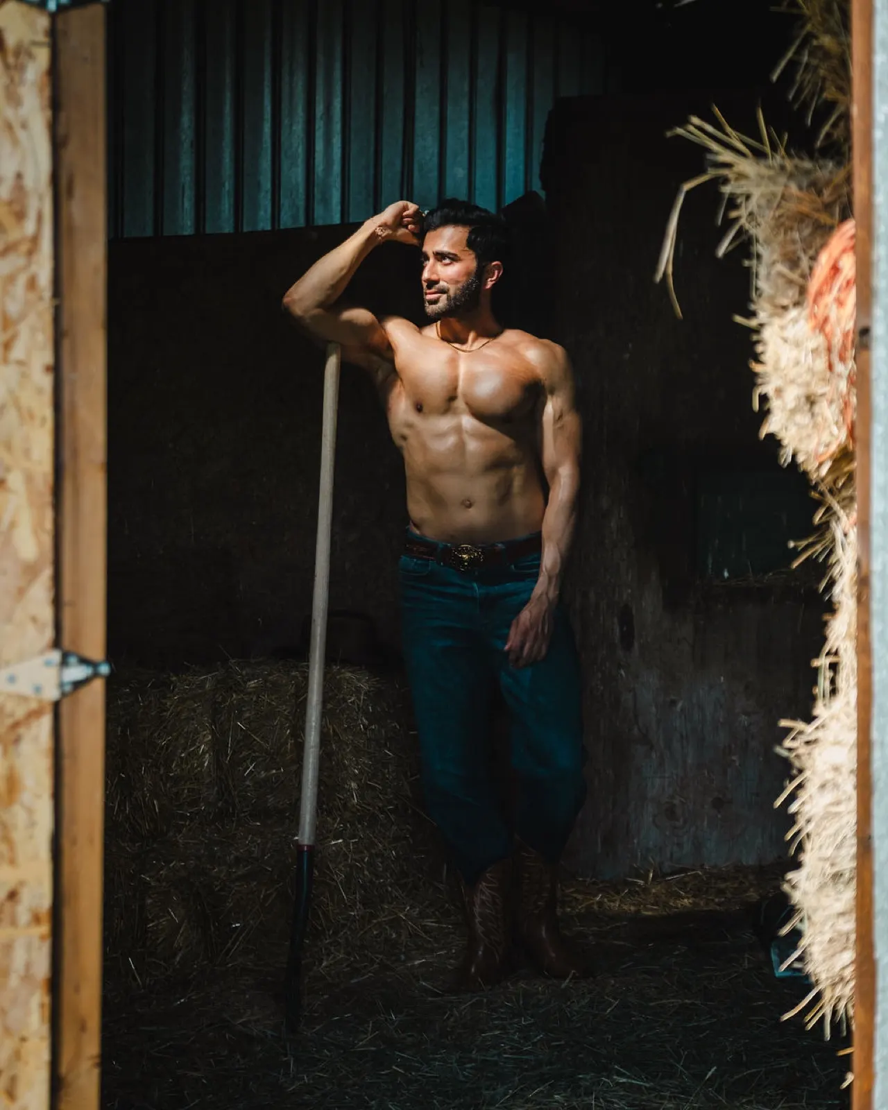 Hemel stands inside a dark stable holding a tool, lit by a warm shaft of sunlight.