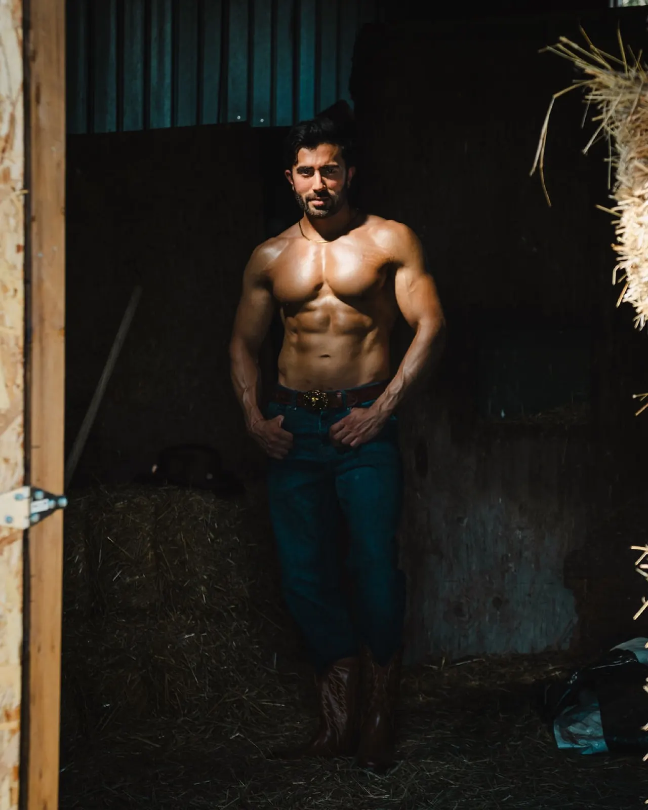 Hemel stands in a dim stable interior with dramatic light emphasizing his physique and western styling.