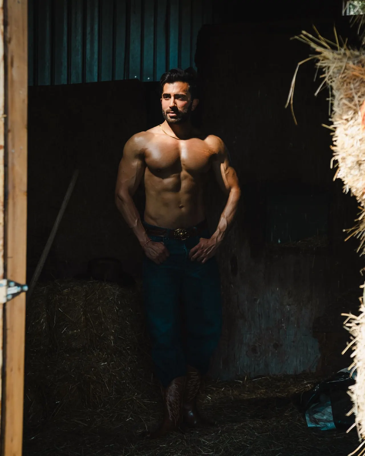 Hemel looks to the side in a shadowed stable, styled with denim, boots, and a western belt.
