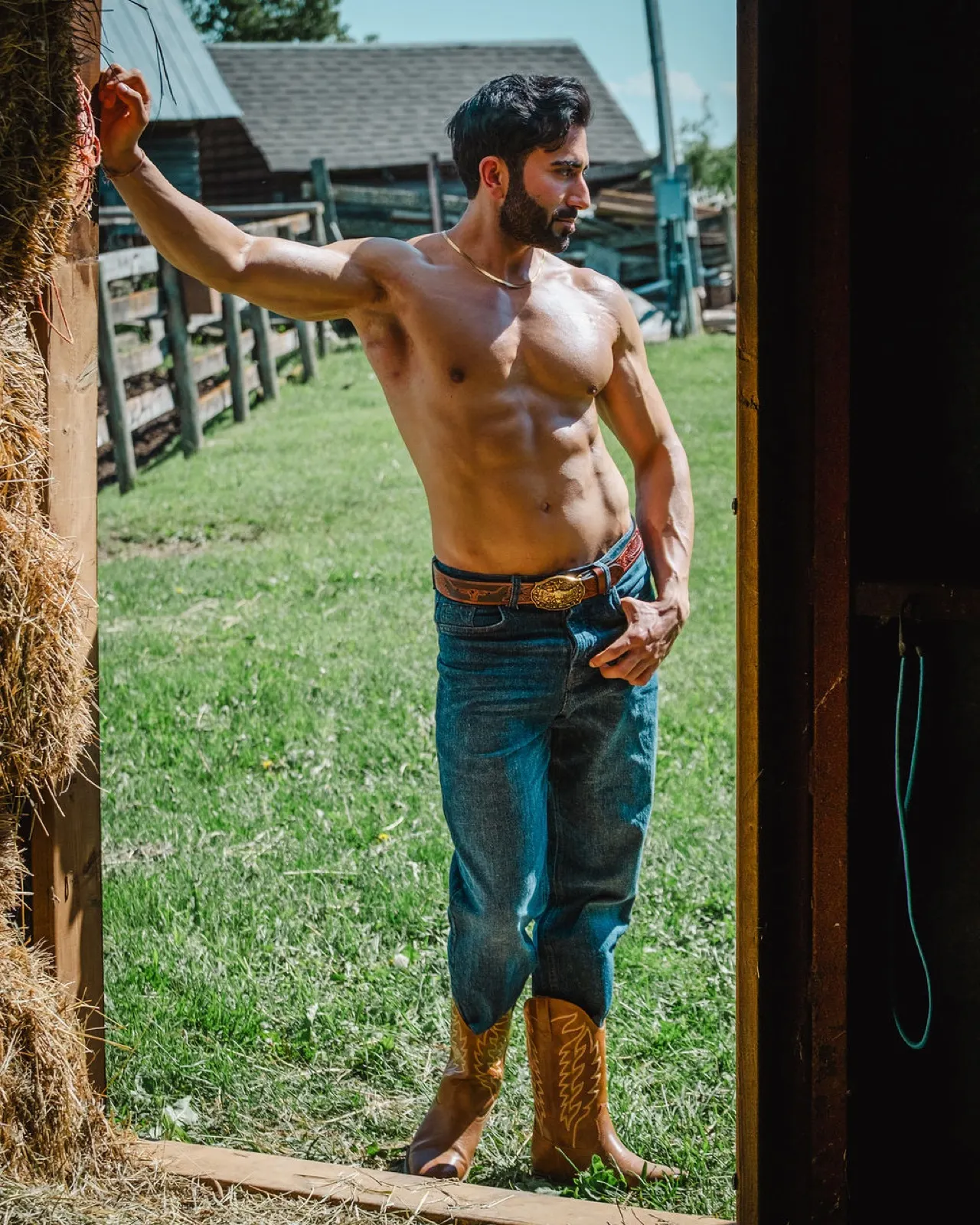 Hemel stands in a stable doorway with sunlight on his torso, framed by hay and ranch buildings.