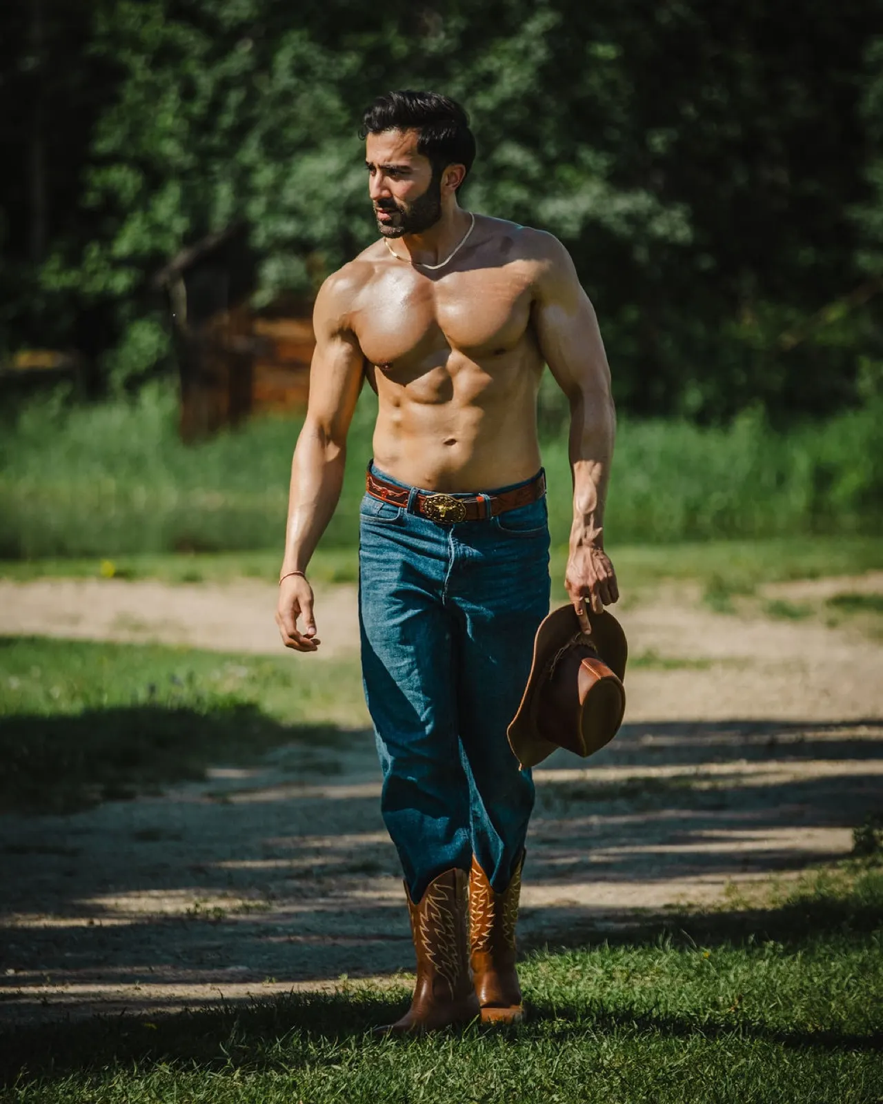 Hemel walks outdoors through a ranch setting, shirtless in jeans and cowboy boots while carrying a brown hat.