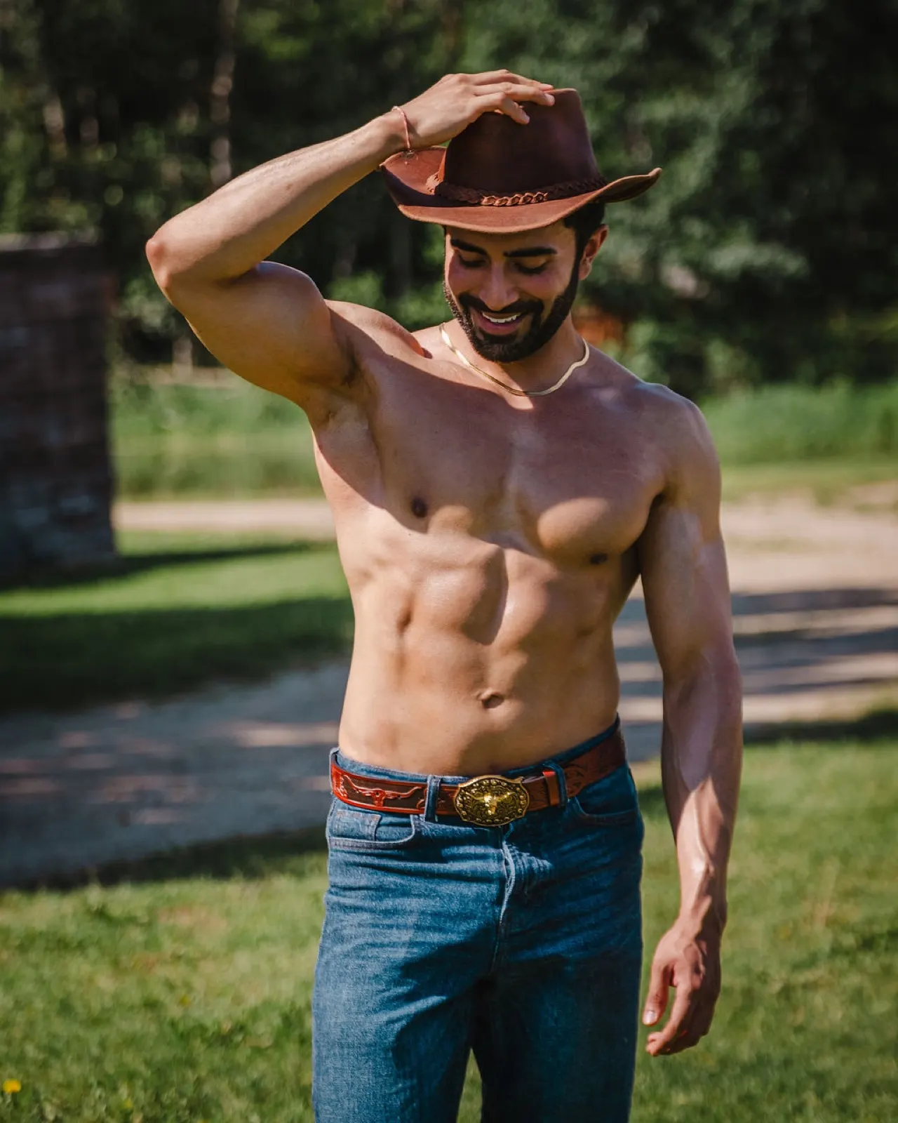 Hemel smiles while adjusting a brown cowboy hat outdoors in a relaxed ranch portrait.