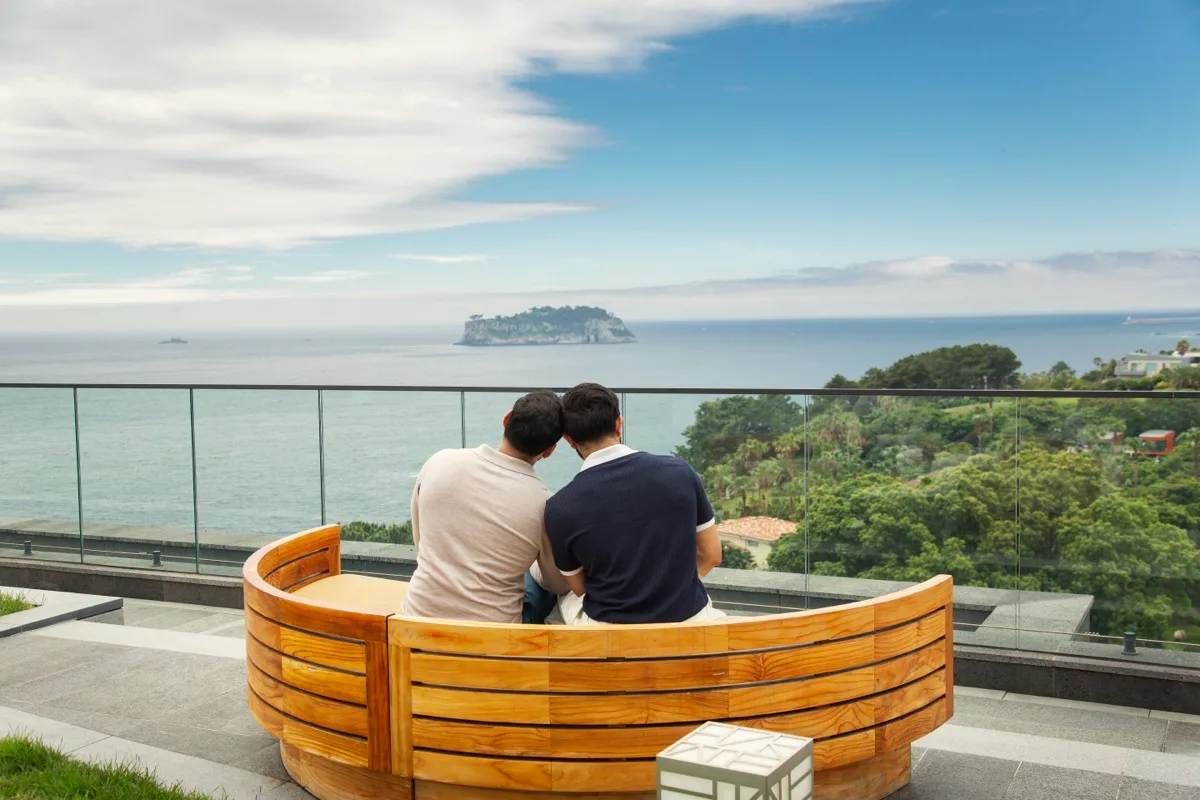 Hemel and Sundeep sitting close together on a rooftop overlooking the ocean in Jeju