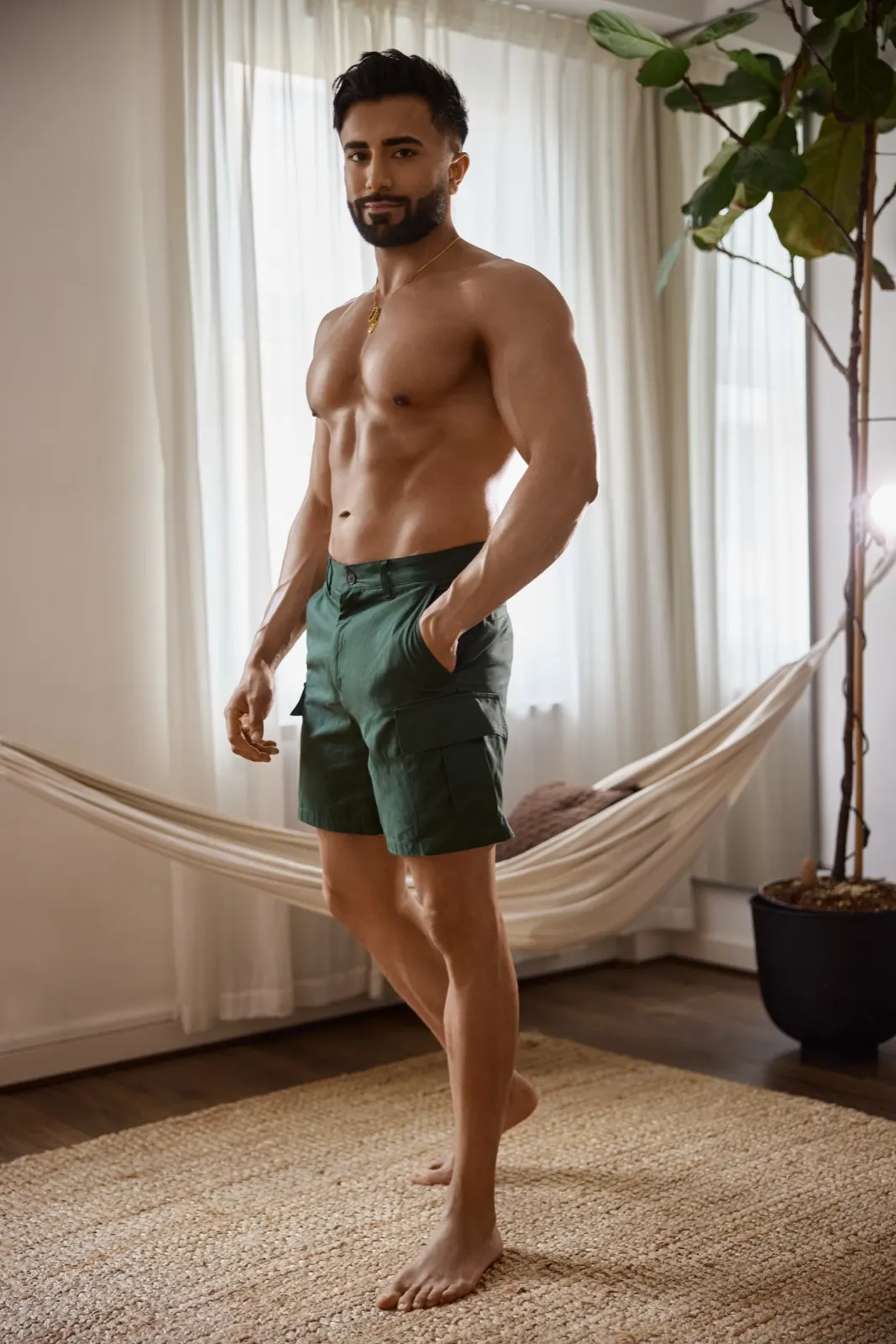 Hemel stands barefoot in green shorts near a hammock and large plant in a bright indoor lifestyle setting.