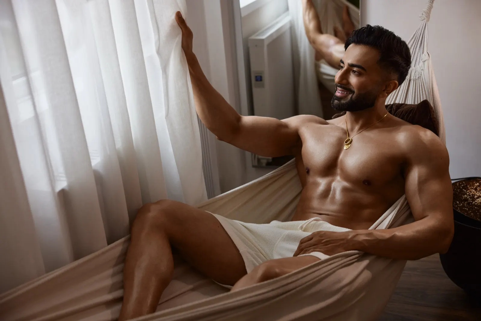 Hemel lounges in a cream hammock by sheer curtains, wearing white shorts in a calm lifestyle scene.
