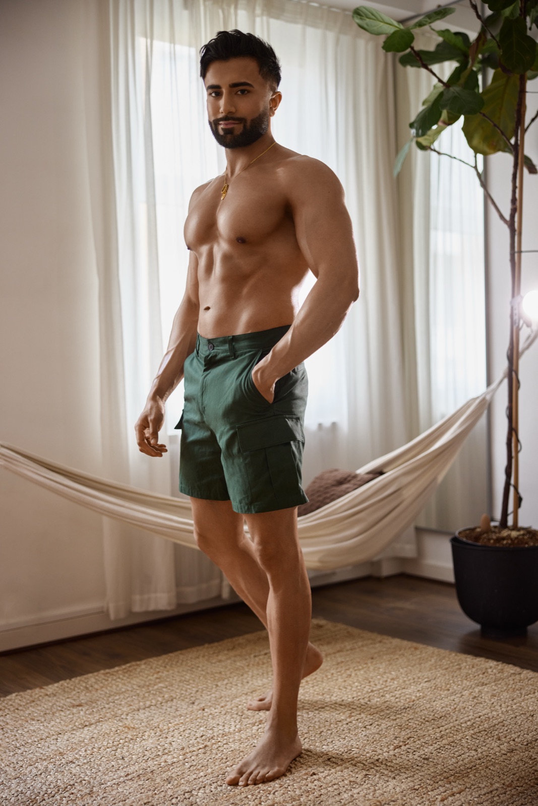 Hemel stands barefoot in green shorts near a hammock and large plant in a bright indoor lifestyle setting.