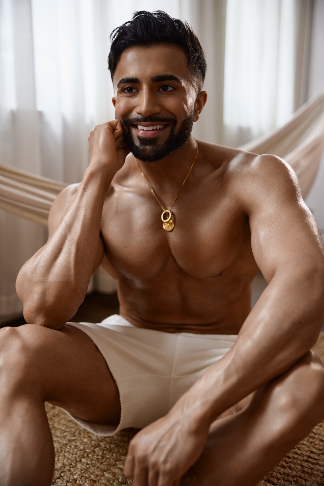 Hemel sits casually in white shorts, smiling beside a hammock and soft curtains in natural light.