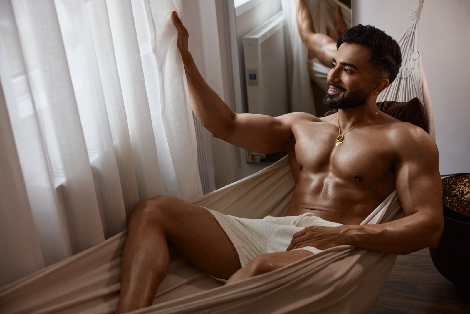 Hemel lounges in a cream hammock by sheer curtains, wearing white shorts in a calm lifestyle scene.