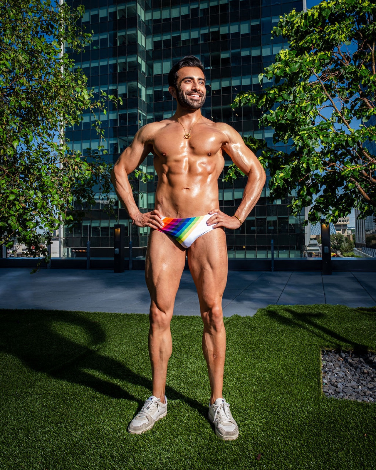 Hemel stands with hands on hips in rainbow swim briefs, surrounded by greenery and reflective city architecture.