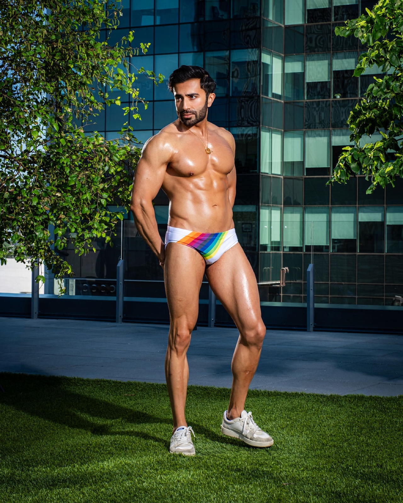 Hemel poses in profile with a focused expression, wearing rainbow swim briefs in front of a glass building.